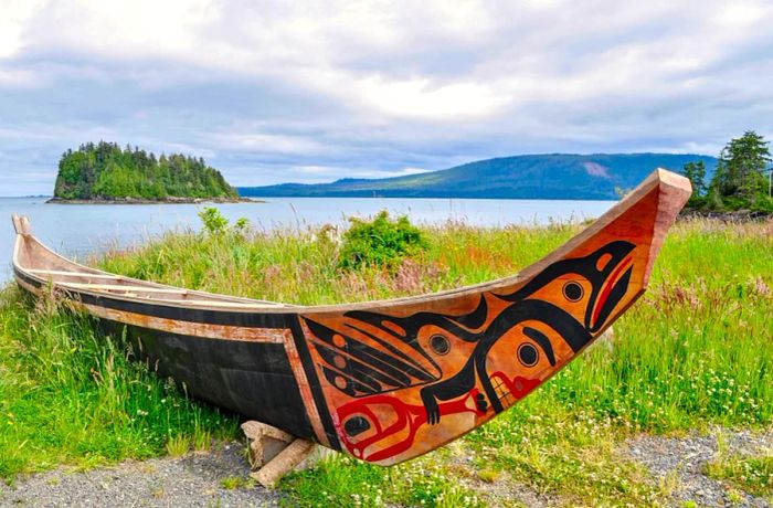 An Aboriginal canoe resting on a grassy bank, with the Pacific Ocean and islands visible in the background on a cloudy day.