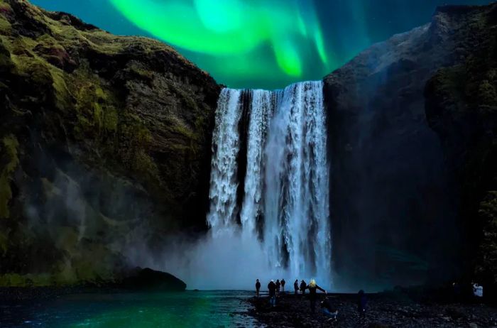 The green northern lights shimmer behind the Skógafoss waterfall in Iceland, with a few visitors admiring the scene in the foreground.