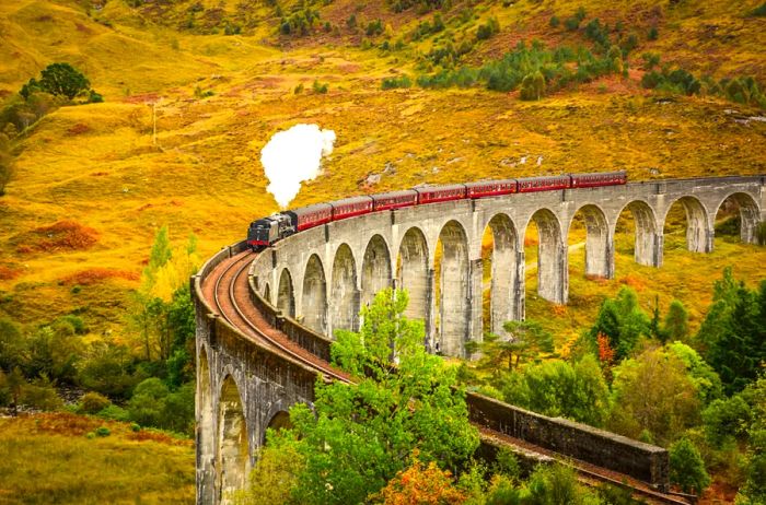 A red train travels atop a curved aqueduct surrounded by golden fields in Scotland