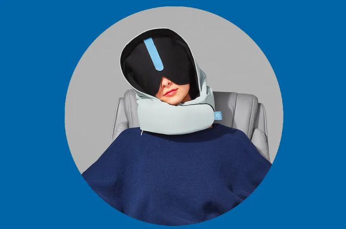 A woman using a gray Pluto Pod travel pillow paired with a large, dark eye mask