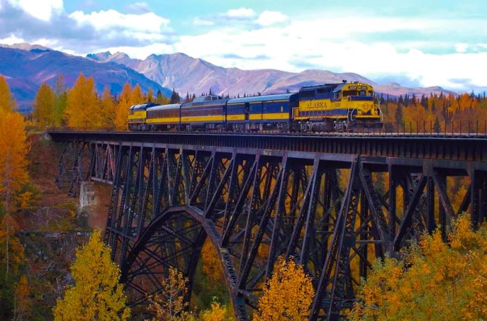 Alaska railcars traverse a trestle bridge with towering mountains in the background