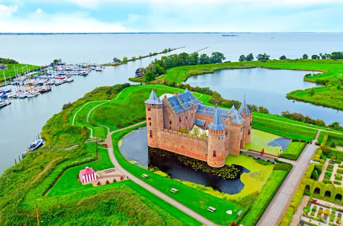 Muiderslot, a medieval castle surrounded by a moat and lush green fields, overlooks the ocean with a line of boats docked to its left.