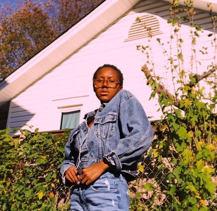 D.C. singer-songwriter Neffy stands in front of plants and the White House, wearing a denim jacket and jeans.