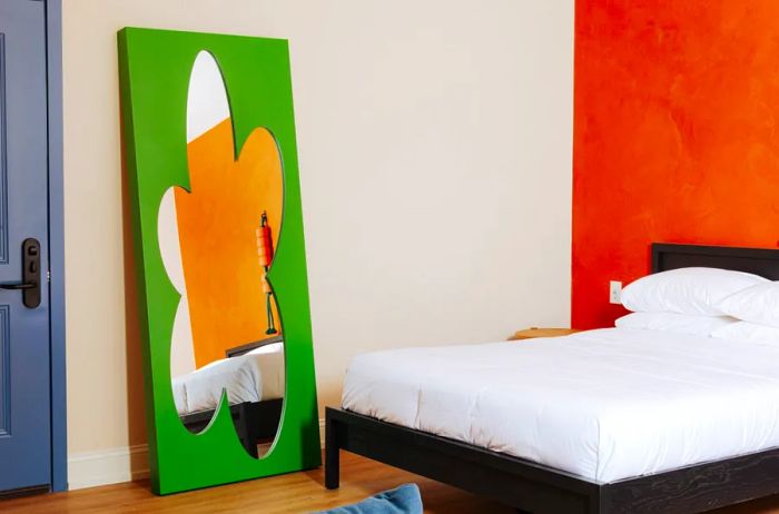 A guest room at Yowie featuring an orange wall behind the bed and a large green floral mirror leaning against a beige wall.