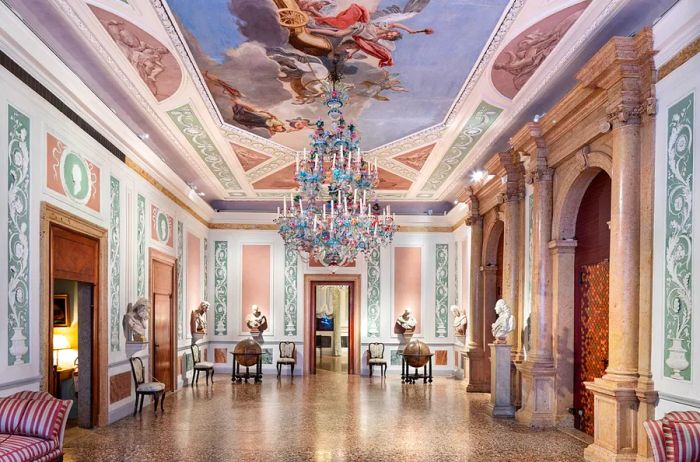 The interior corridor of Casa Museo Querini Stampalia features elegant marble, a grand glass chandelier, small busts, and a beautifully painted high ceiling.