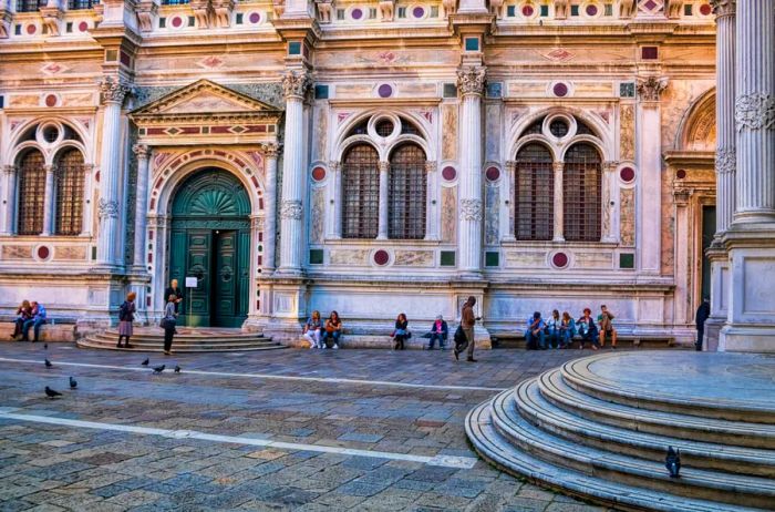 The exterior of Scuola Grande di San Rocco features arched windows and doorways, with about a dozen people seated on its steps.