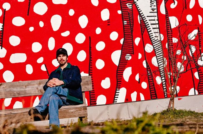 Conrad Benner sitting outdoors on a wooden bench in front of a vibrant red, white, and black mural.