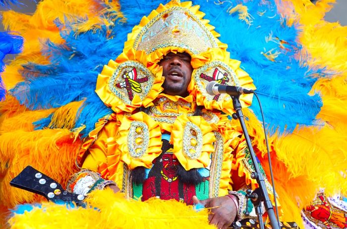 A Black Masking Indian adorned in a stunning gold and silver headdress and outfit, surrounded by vibrant blue and yellow feathers, stands before a microphone at Jazz Fest.