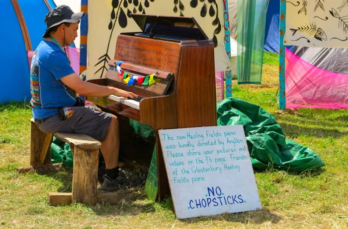 A man playing the piano on a rustic wooden bench outdoors at the Glastonbury Festival