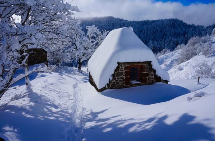 A quaint stone cottage blanketed with a thick layer of snow on its roof, nestled amidst snowy hills and surrounded by snow-covered trees.