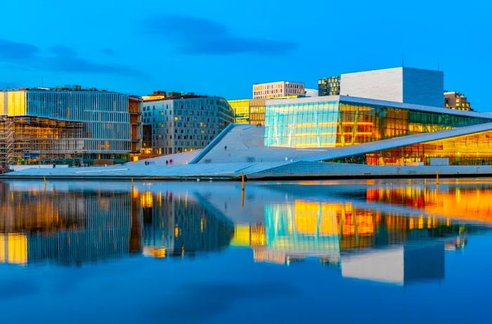 The Oslo Opera House, Norway, illuminated at twilight, with its reflection shimmering in the water and boxy buildings in the background.