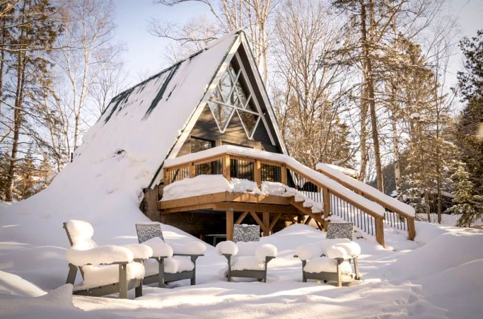 An A-frame cabin adorned with snow on its roof and the railings of its deck and staircase, featuring four chairs in the foreground, blanketed by a thick layer of snow.