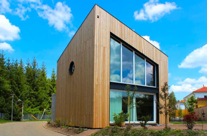 A cube-shaped apartment clad in wood features a large window that nearly spans the entire front wall, with evergreen trees in the backdrop.