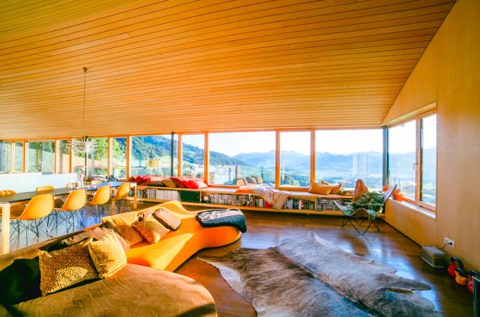 A spacious living area featuring an animal skin rug, an orange sofa adorned with pillows, a long dining table surrounded by eight orange chairs, and a large window showcasing views of a valley framed by rocky mountains.