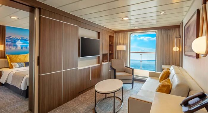 A suite on the Aurora Expeditions' Sylvia Earle vessel features a bedroom in the background and a separate living area with a couch, TV, and ocean-view balcony in the foreground.