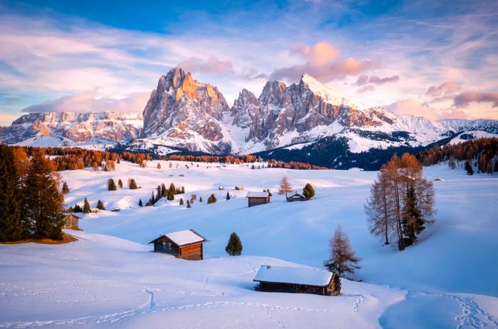 Charming cabins dot a snowy landscape in Italy's Dolomites, framed by the majestic, snow-covered rocky mountains in the background.