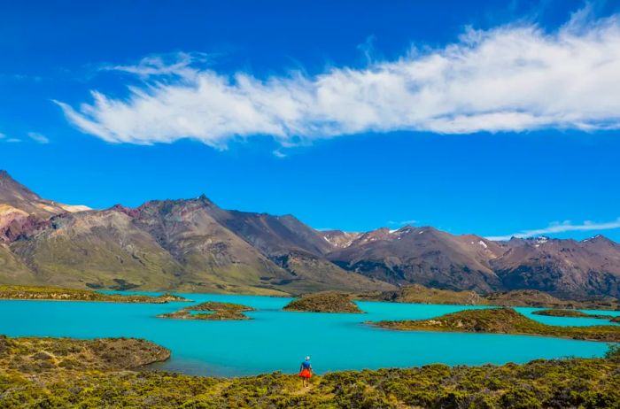 A hiker stands in the foreground against a backdrop of icy blue lake and rugged gray mountains in Perito Moreno National Park, Patagonia.