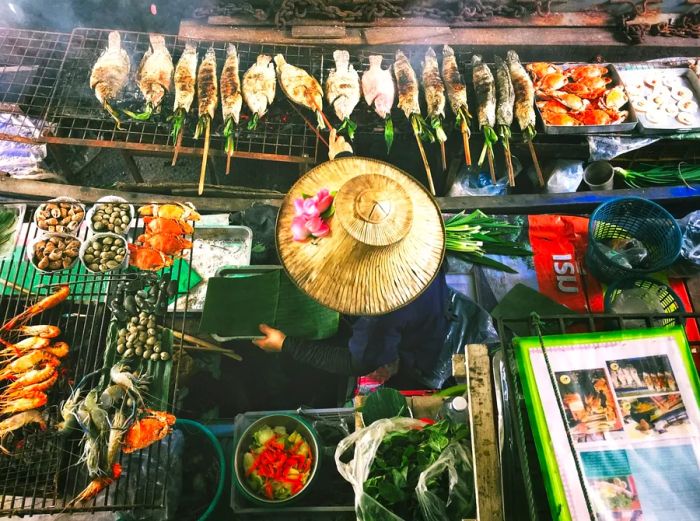 An aerial view of a person in Thailand, wearing a wide-brimmed hat, surrounded by grilled fish and seafood.