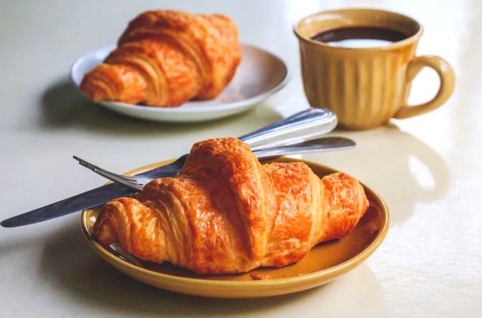 Two French croissants on plates, accompanied by a cup of black coffee in the background.