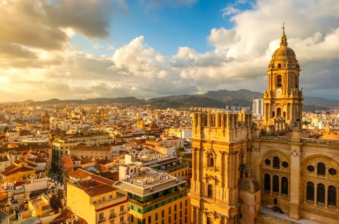 An aerial view of Málaga, Spain, featuring a cathedral in the foreground surrounded by a mix of historic and modern buildings, with a backdrop of clouds and blue skies.