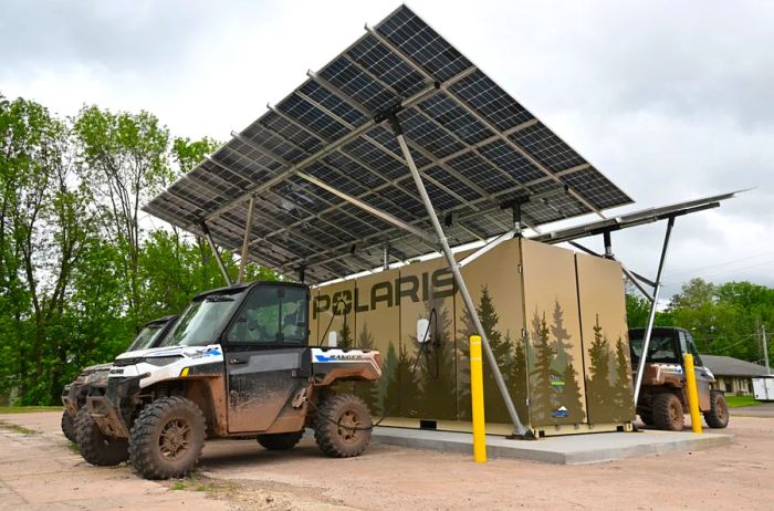 Several mud-splattered Polaris vehicles are parked at a charging station resembling a giant cardboard box adorned with tree designs.