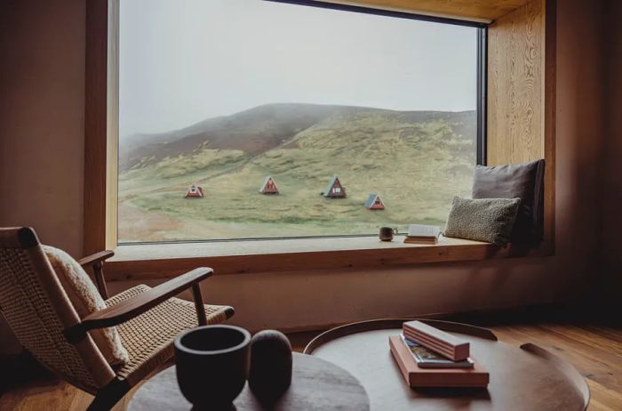 An armchair positioned by a large picture window, offering a view of several A-frame cabins on a verdant hillside.