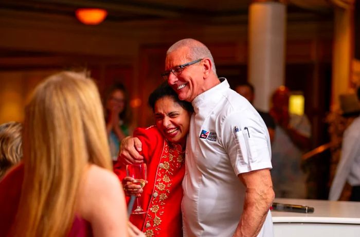 Chefs Maneet Chauhan and Robert Irvine share a friendly moment, smiling in a side hug, while Chauhan enjoys a glass of white wine, surrounded by cruise passengers.