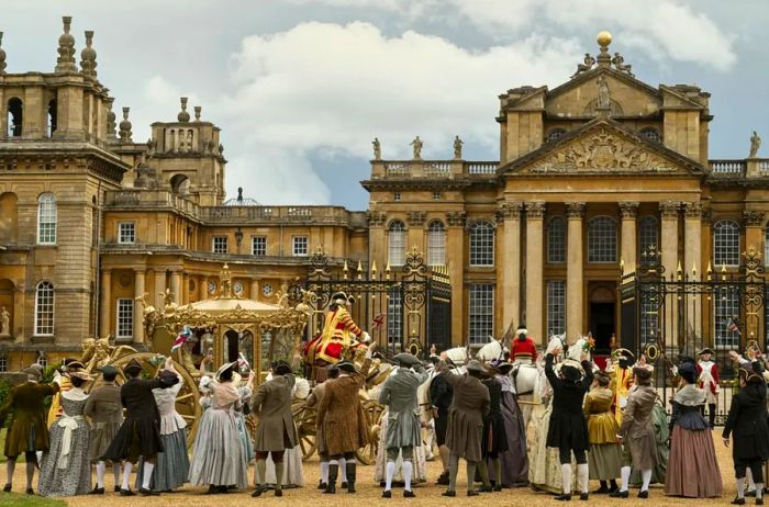 A group of individuals dressed in period costumes stands outside the fictional Buckingham House, with the impressive exterior of Blenheim Palace visible in the background.