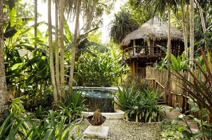 A treehouse featuring a grass roof, a small pool below, and lush greenery all around.