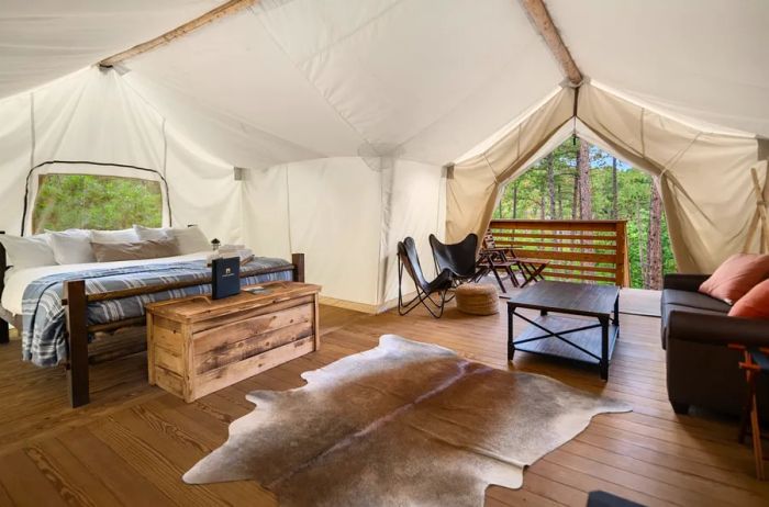 A sDinogoi-style tent featuring a wooden bed, a cozy couch, a deerskin rug on the floor, a trunk, and two camp chairs, with trees visible outside.