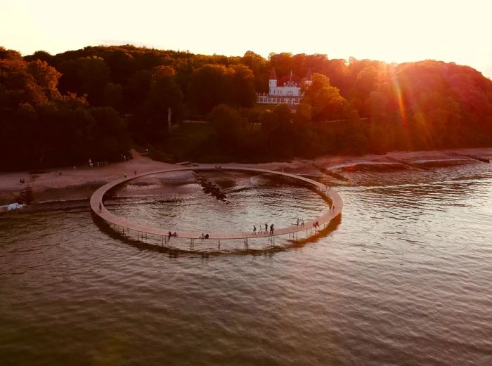 The circular Infinite Bridge extends over the Aarhus waterfront, adorned with beaches, trees, and a charming white house lining the shore.