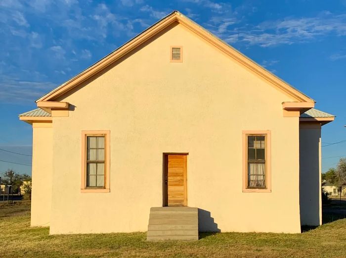 The exterior of the Blackwell School in Marfa, Texas, features a straightforward design with one door, two windows, cream-colored walls, and pink accents.