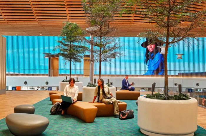 Visitors relax on cushioned benches surrounded by small potted trees, with a large video screen displaying scenes from the Pacific Northwest in the backdrop.