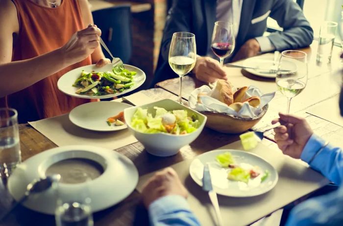 A woman in a dress and two men in suits gathered around a dining table featuring salads, bread, and wine