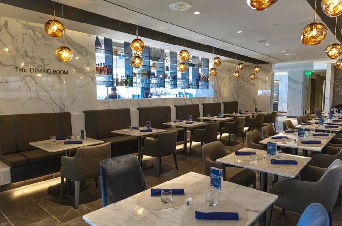 A series of marble-topped tables and dining chairs with a bar in the background, illuminated by amber globe lights at the United Polaris lounge in Houston