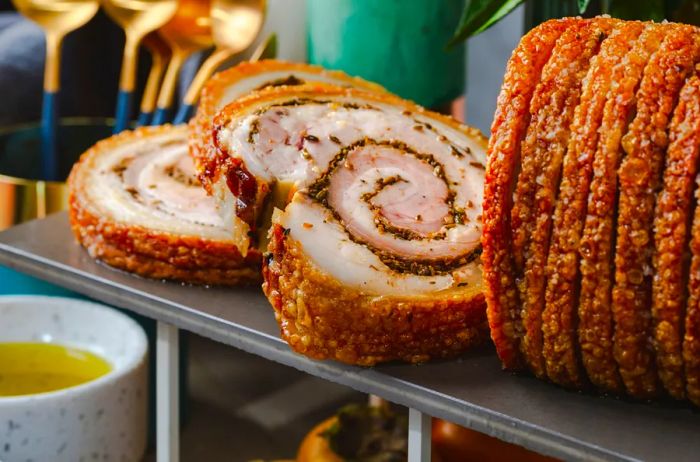 A roll of porchetta, featuring rolled pork with three thick slices cut from the log. The exterior is crispy and golden brown, resting on a grey table.