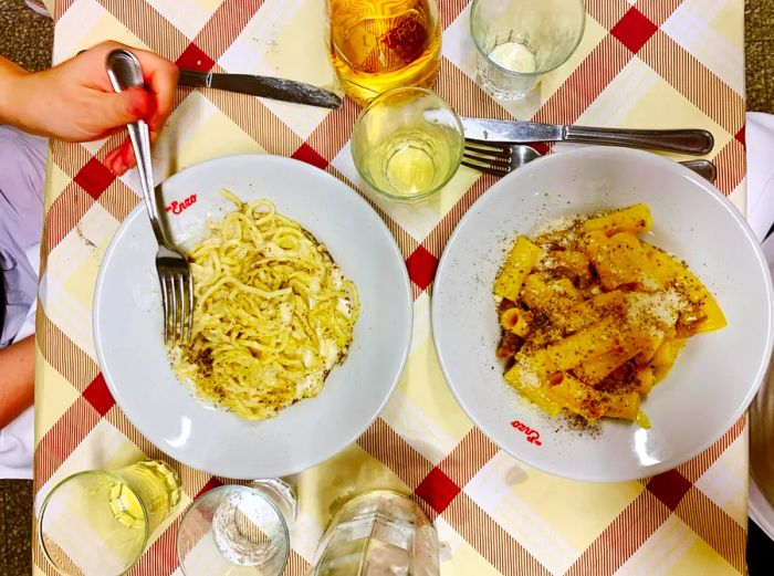 An overhead view of two bowls of pasta on a table adorned with a yellow and red checkered tablecloth. On the left, a bowl of cacio e pepe with a woman reaching for it with her fork, and on the right, a bowl of carbonara with a fork and knife resting beside it. Several glasses of drinks surround the dishes.