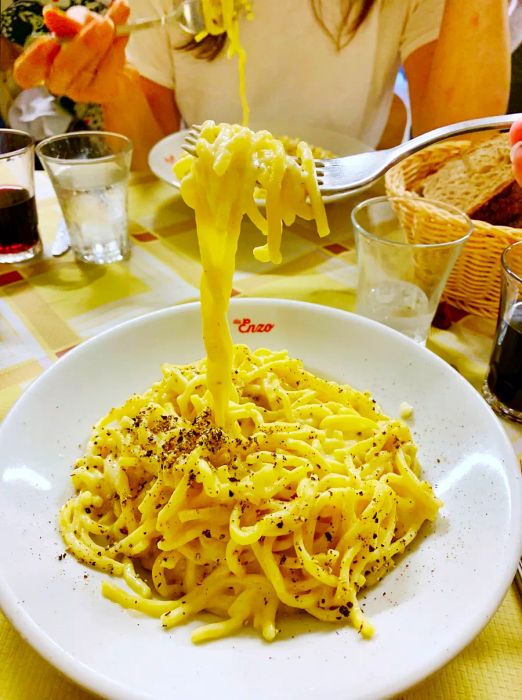 A white bowl brimming with creamy, yellow cacio e pepe, as someone pulls a forkful of pasta from it; this dish is served at Da Enzo al 29.