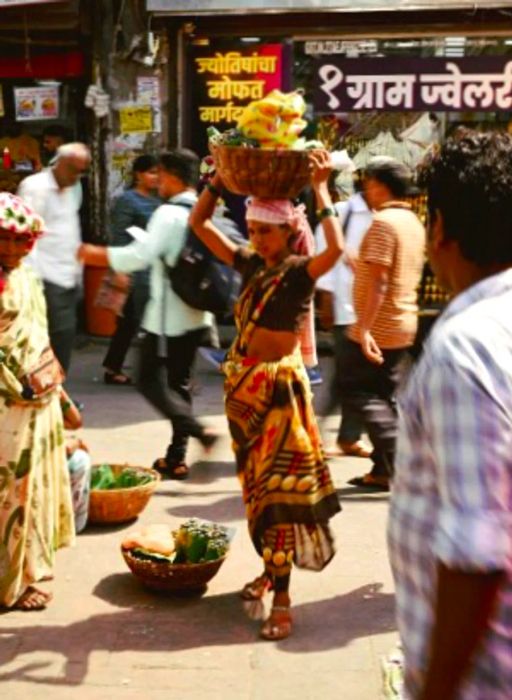 A person carries a basket of fruit balanced on their head while navigating a bustling Mumbai street.