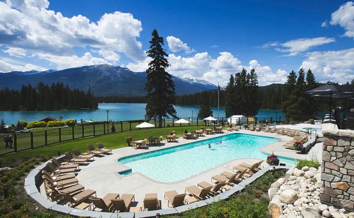 A spacious rectangular outdoor pool surrounded by unoccupied tan lounge chairs, adjacent to a lake at Fairmont Jasper Park Lodge.