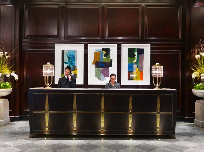 The reception area at Hotel Georgia 274 features two staff members and is adorned with three vibrant framed artworks against a dark wood wall.