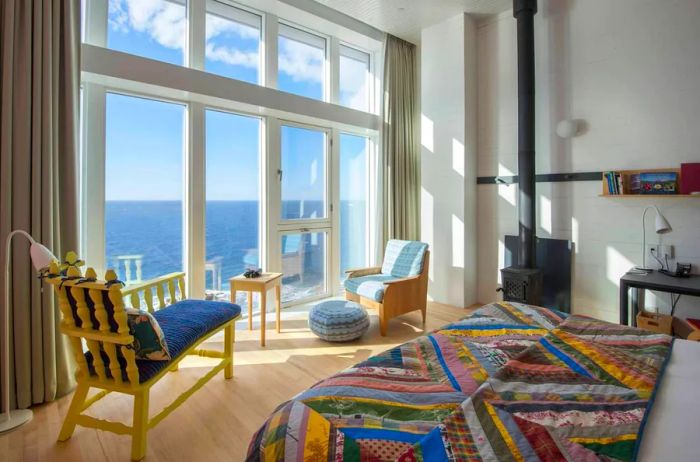 The interior of a bright guest room at Fogo Island Inn features a colorful quilt on the bed and floor-to-ceiling windows that overlook the water.