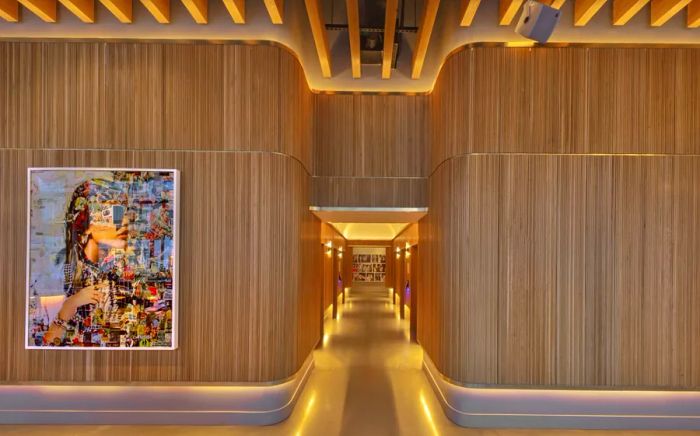 The lobby of the Virgin Hotel in NYC features a hallway leading to the elevators, illuminated by lights along the lower wall reminiscent of an airplane aisle, complemented by wood-paneled walls.