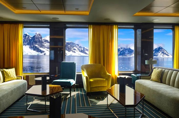 A room featuring two sofas and armchairs, adorned with yellow floor-to-ceiling drapes; four large windows provide views of snowy peaks.