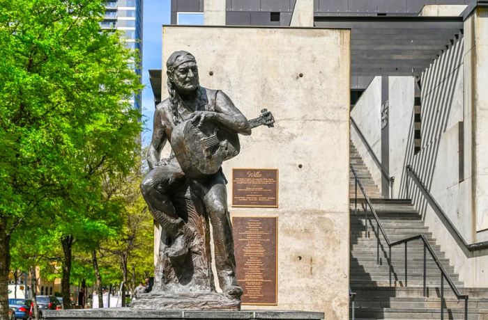 A statue of Willie Nelson with long braids and a guitar in front of a cement wall featuring a bronze plaque