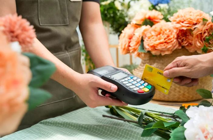 A customer using a credit card reader held by a shopkeeper, with fresh flowers adorning the counter and background.