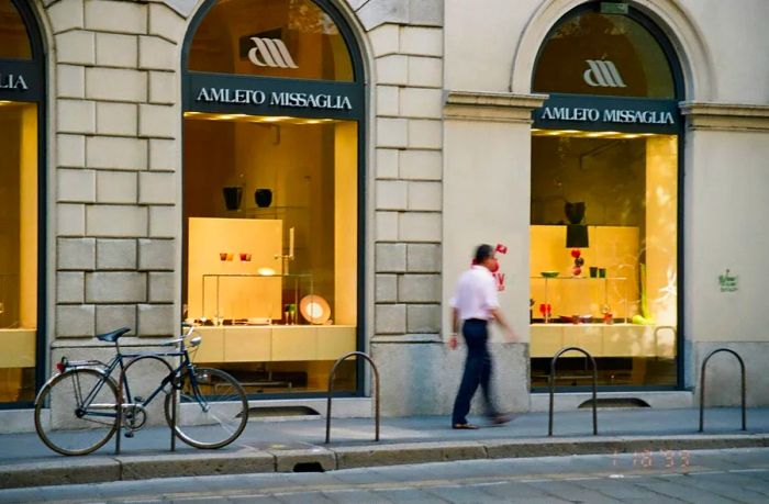 A person strolls past a pair of arched shop windows, with a bicycle parked on the sidewalk.