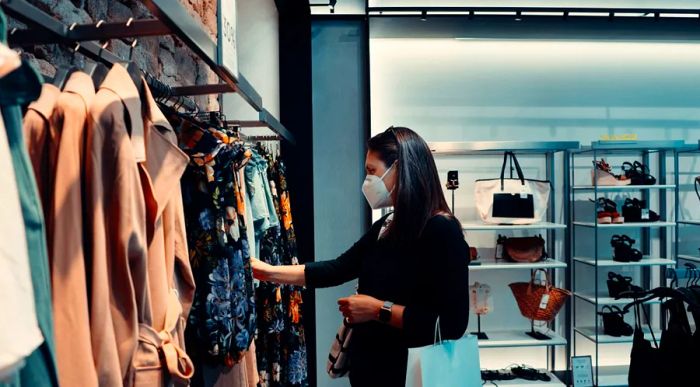 A woman in a mask browsing through clothing racks, with shelves of bags and shoes behind her