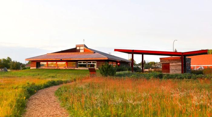 A footpath meandering through a prairie at sunrise, with a low wooden building visible in the background.