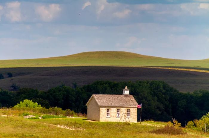 A quaint stone one-room schoolhouse perched on a grassy hill, with a larger hill looming in the distance.